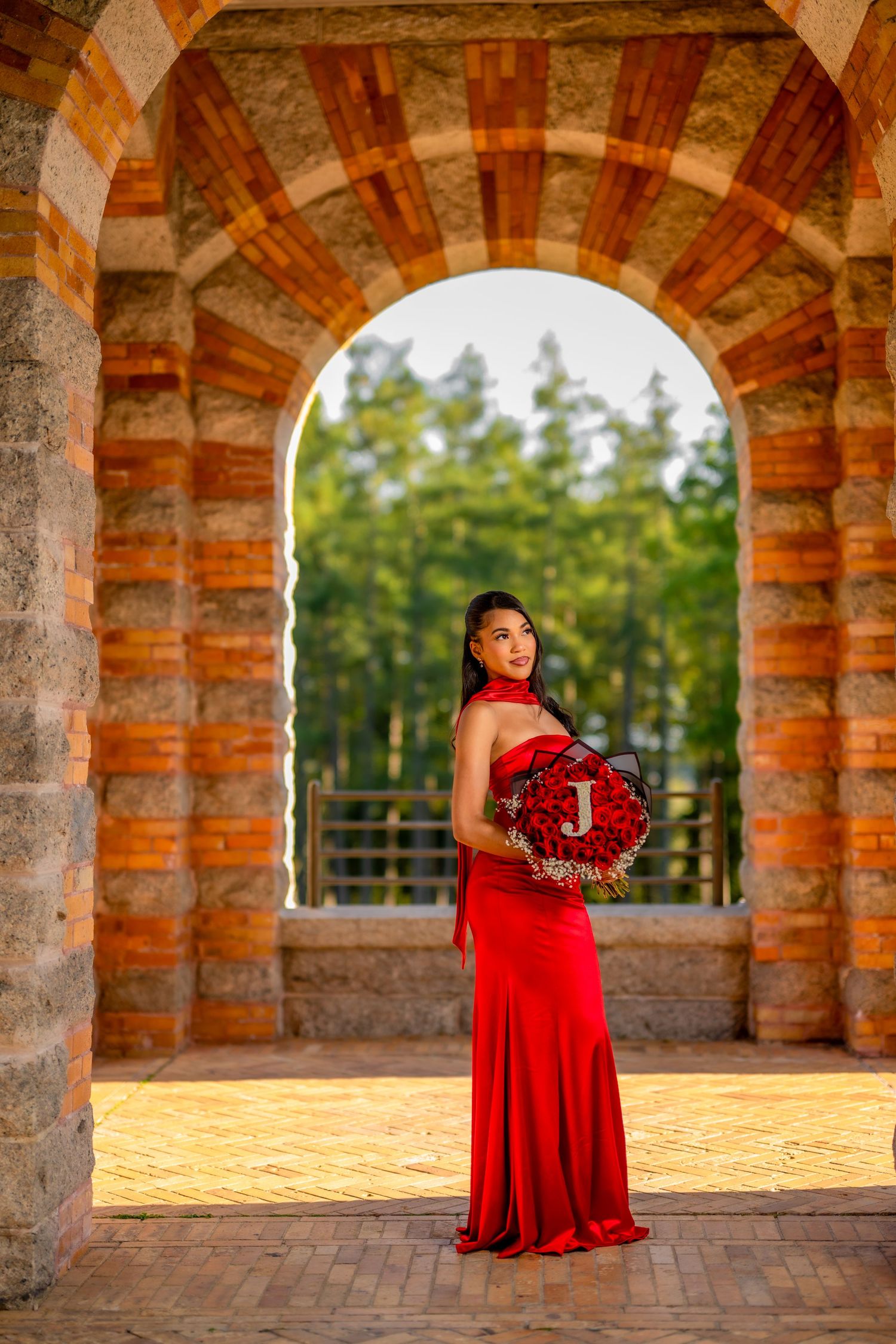 Woman in red gown holds floral bouquet under stone arch with garden view behind her.