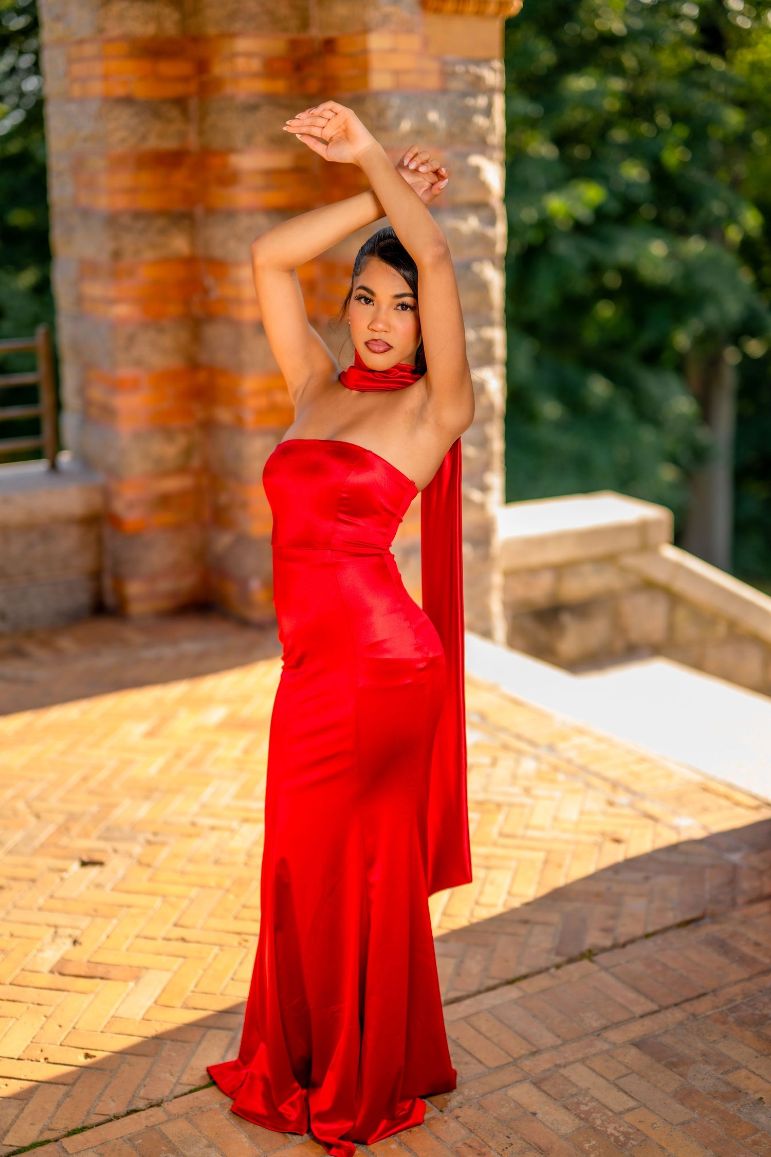 Woman posing elegantly in a flowing red strapless satin gown with halter neck detail outdoors.