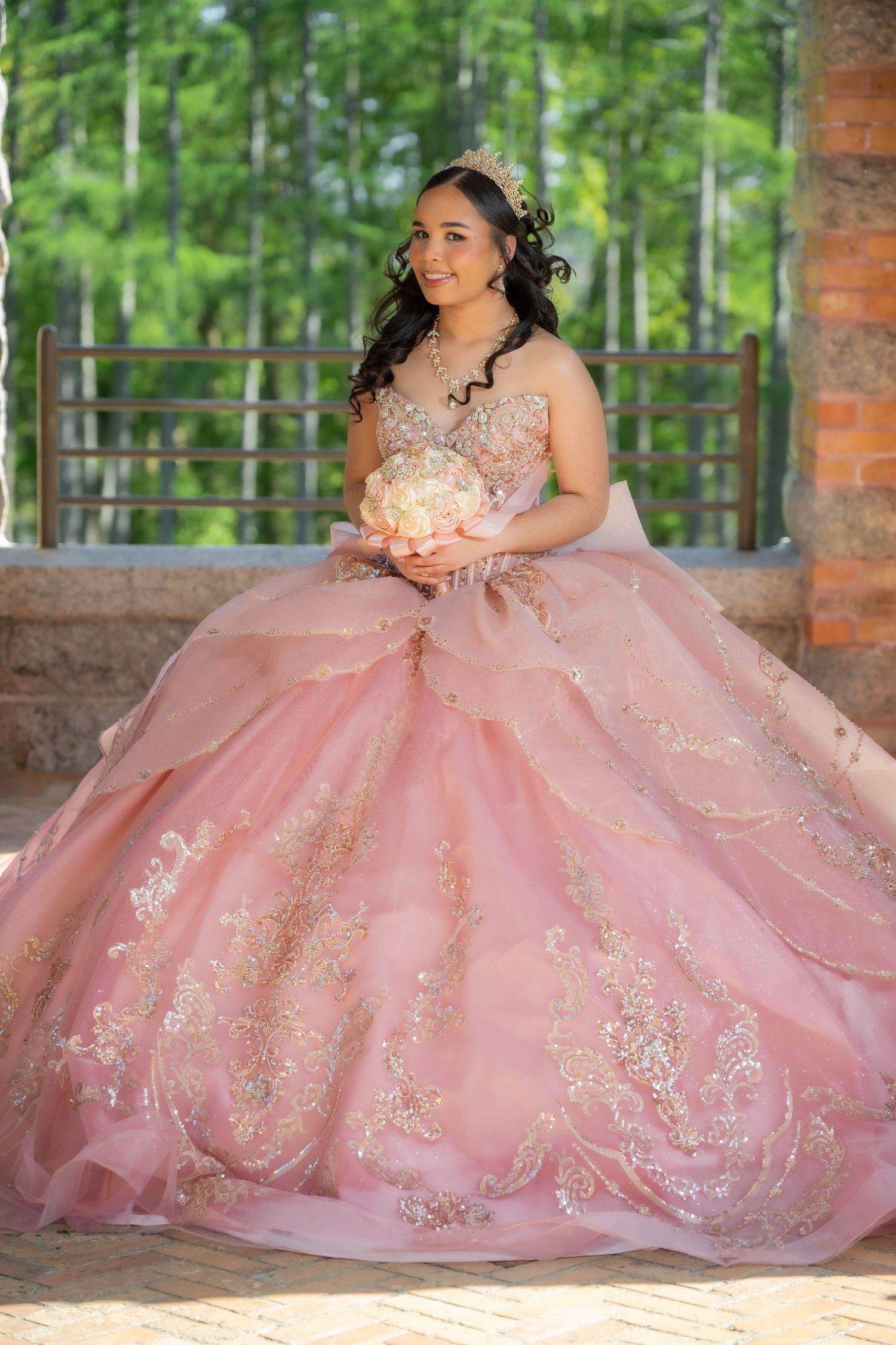 Young woman in pink ballgown with gold embroidery holds bouquet at quinceañera celebration outdoors.