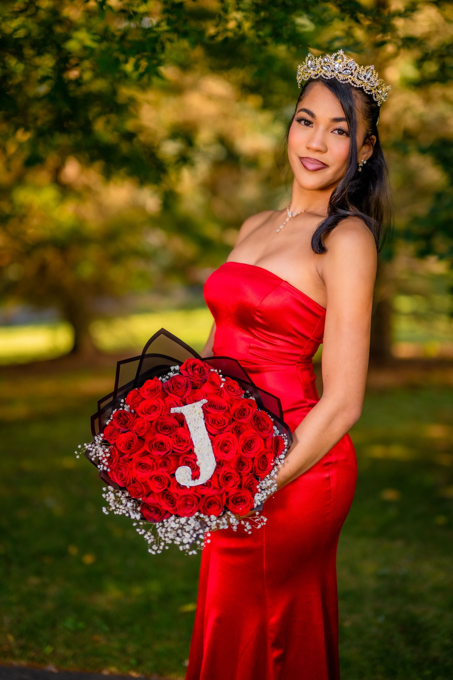 Woman in red strapless gown holding large rose bouquet with J initial wearing tiara crown.