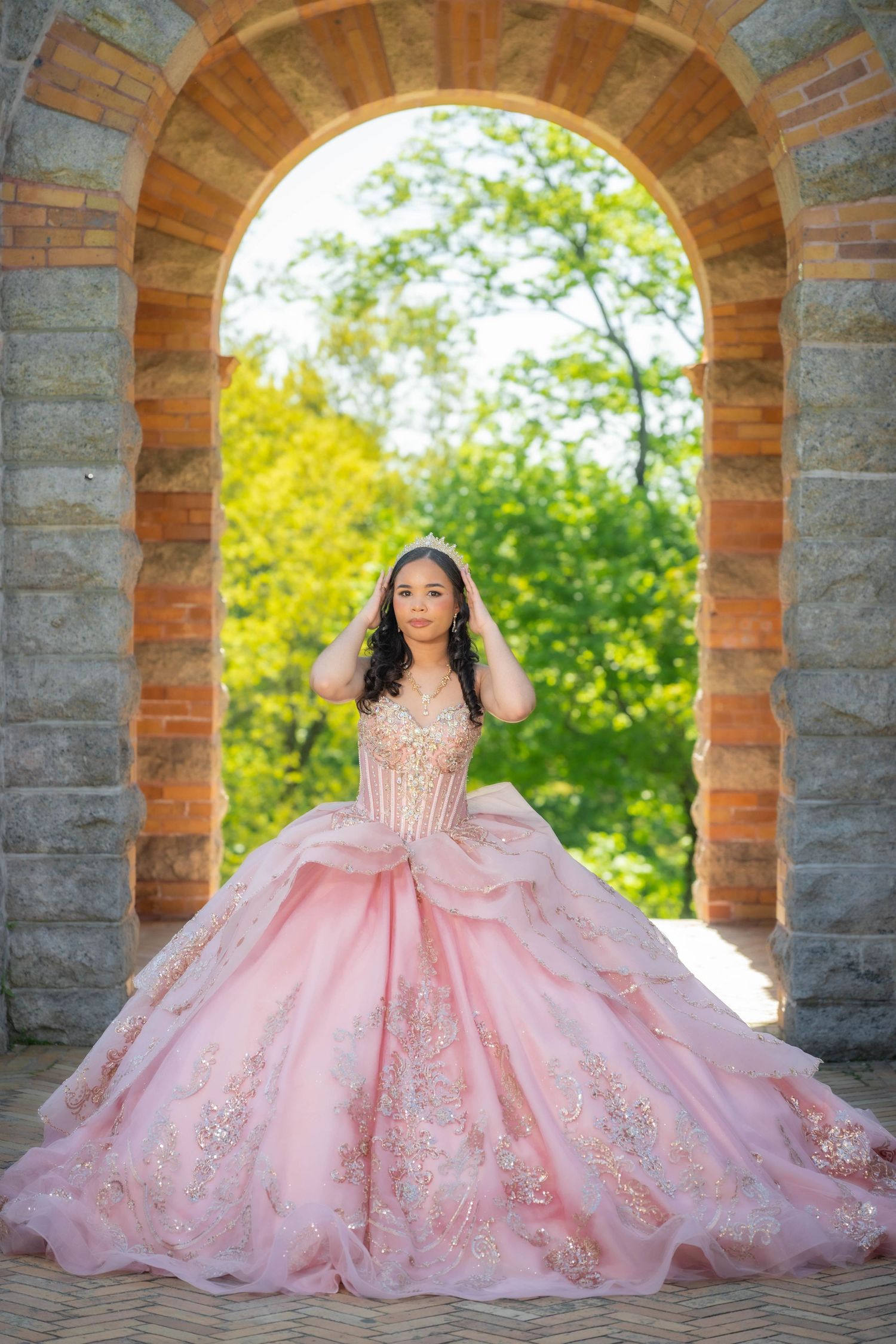 Young woman in pink ballgown with gold embroidery poses under stone arch with lush green trees in background.