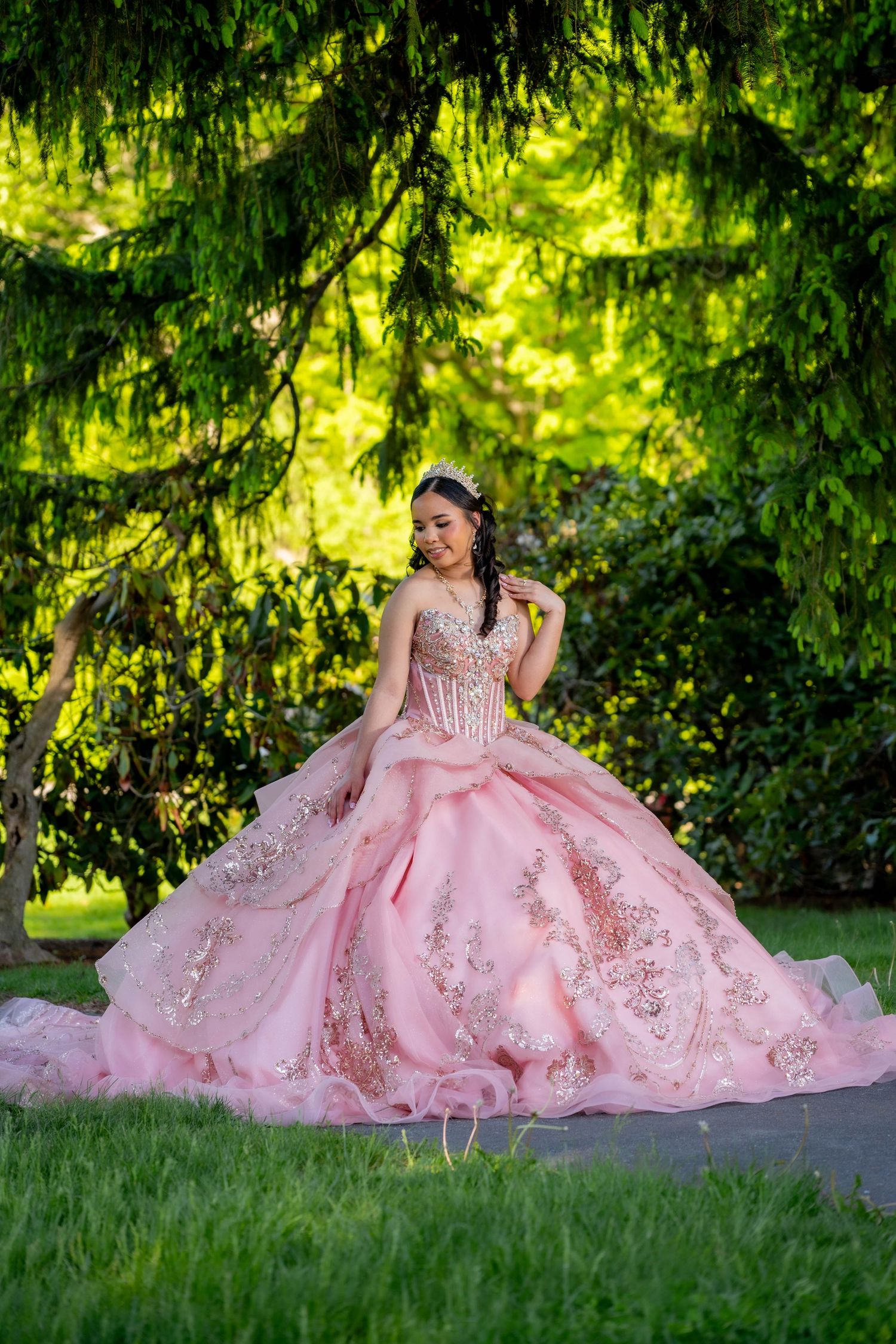 Young woman in pink quinceañera ball gown with gold embroidery poses gracefully outdoors under lush green trees.