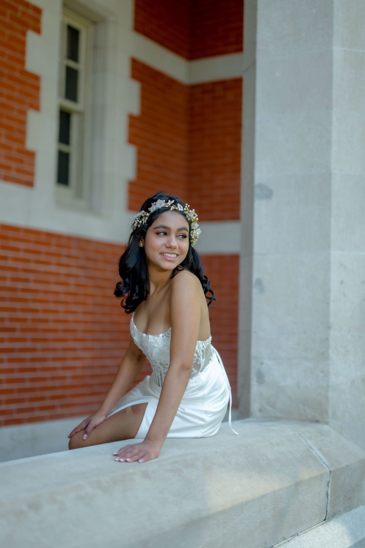 Young woman in white gown and floral crown sits gracefully on stone steps beside a red brick building.