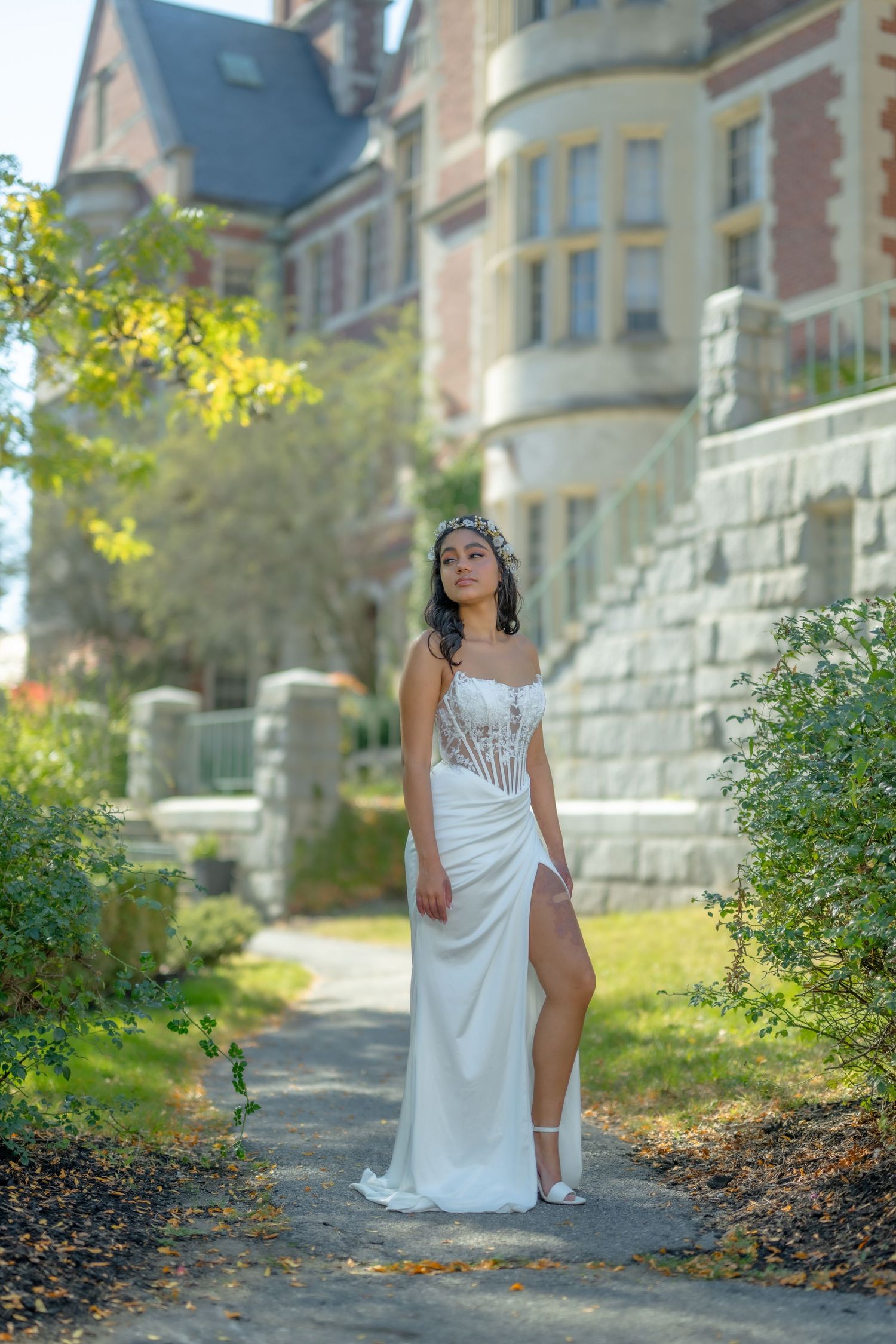 Young woman in elegant white gown with corset detail and crown poses on garden path before historic stone mansion.