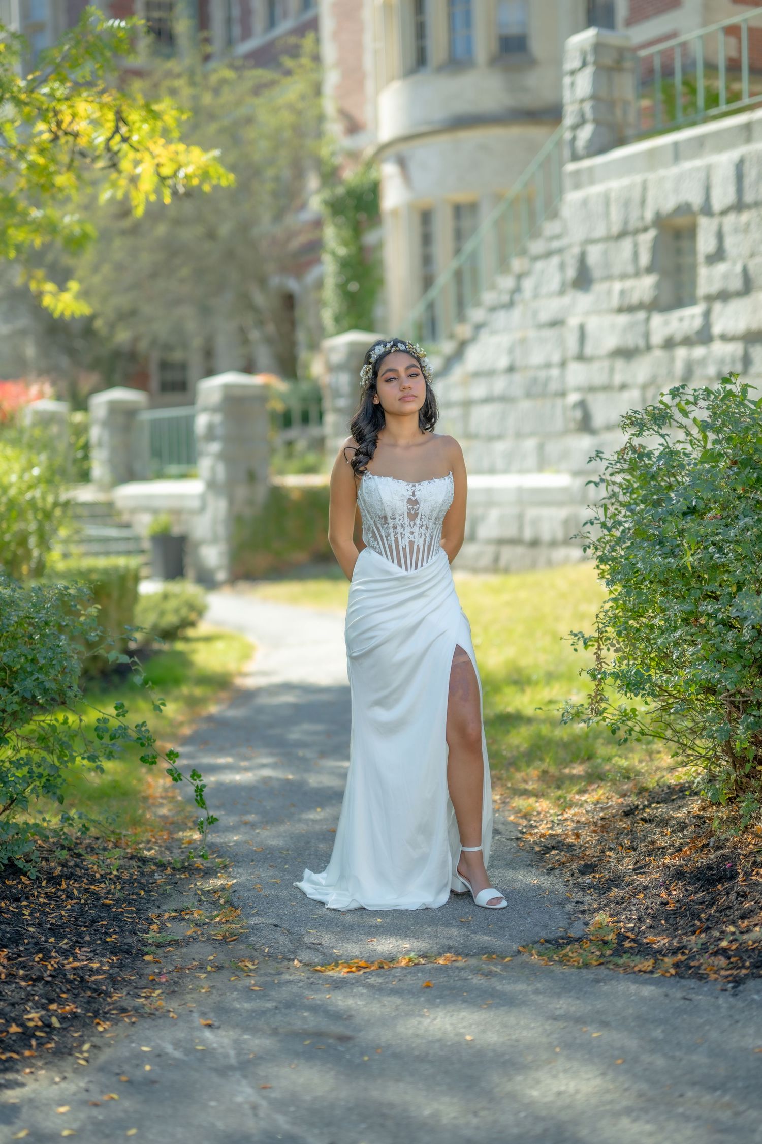 A bride in a white corset gown with a slit poses on a garden path near a stone building.