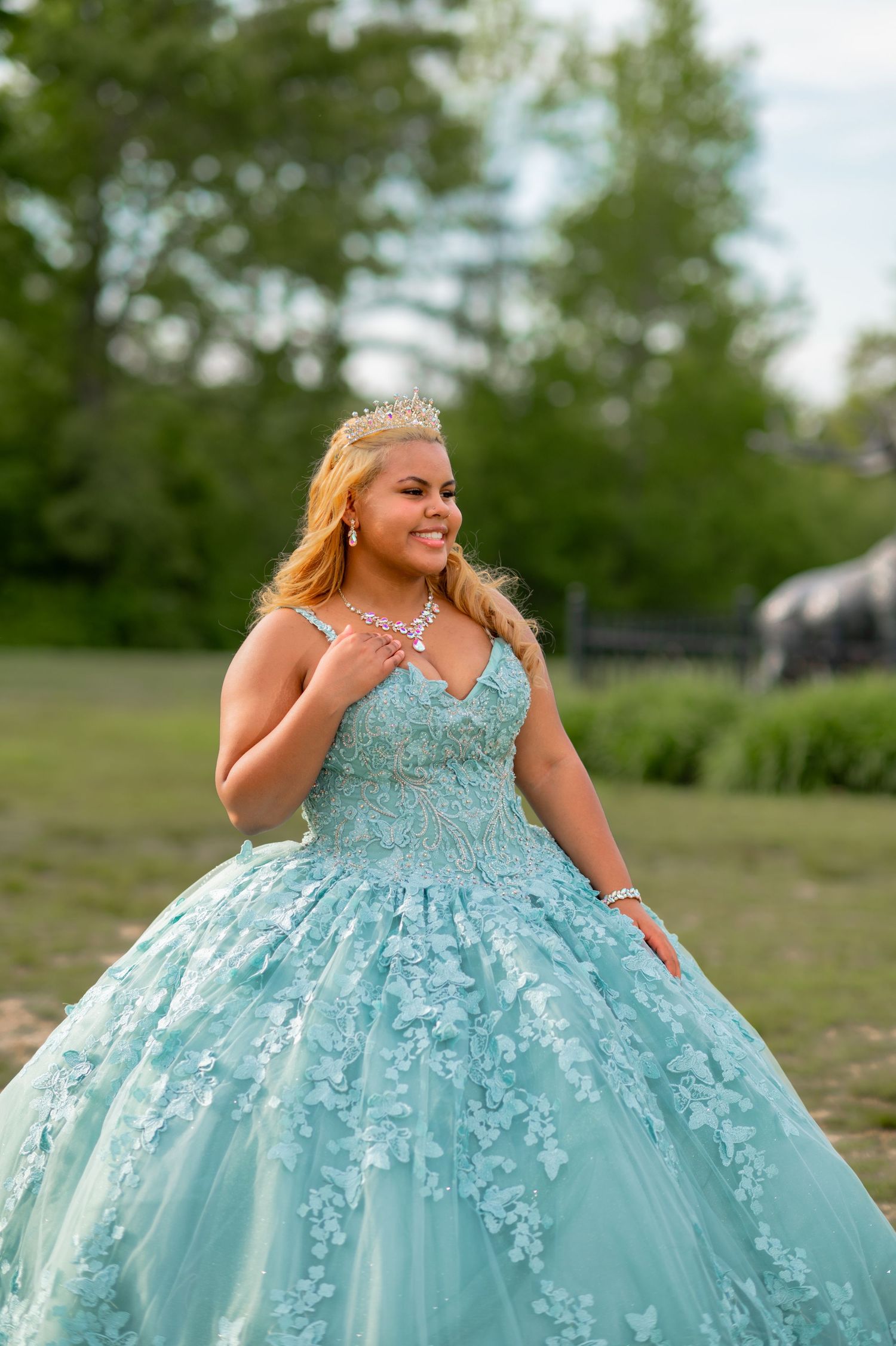 Young woman in a light blue quinceañera ball gown with floral lace, tiara, and jewelry, posing outdoors.