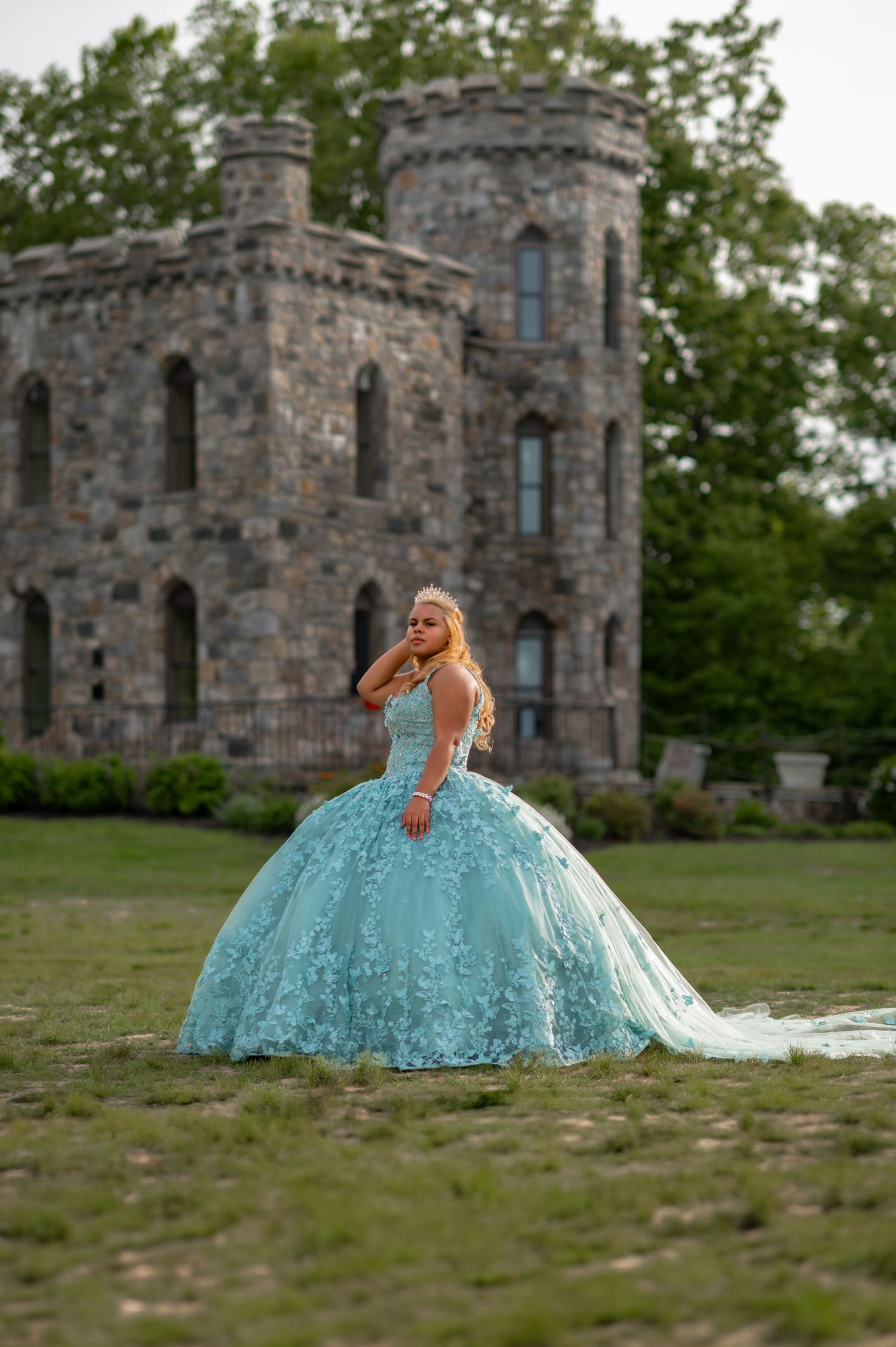 Young woman in a teal ball gown poses in front of a stone castle during a quinceañera photoshoot.