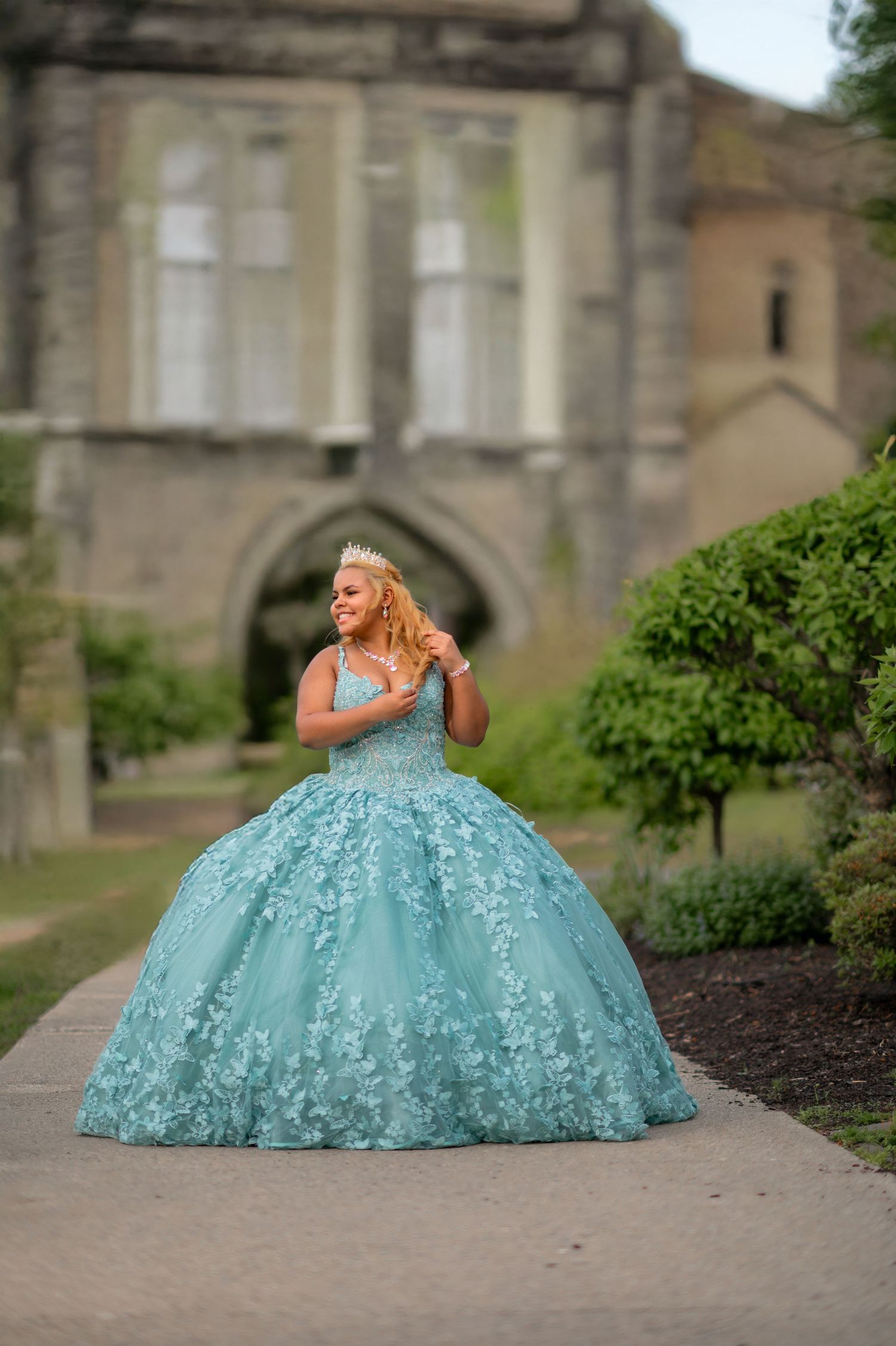 Young girl in a teal quinceañera ball gown with floral lace and tiara poses before a stone castle.
