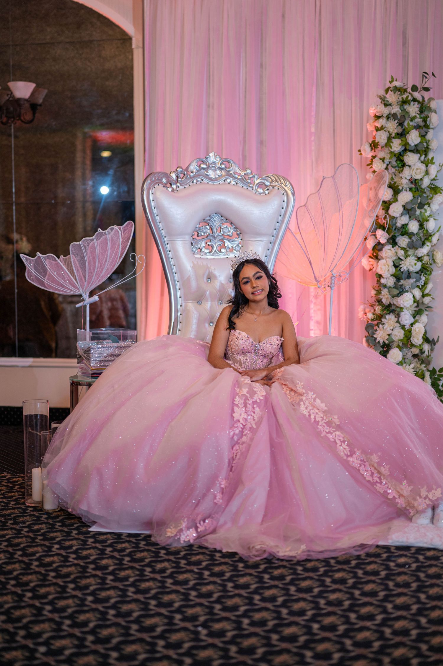 Young woman in pink ball gown quinceañera dress seated on ornate white throne chair with floral backdrop.