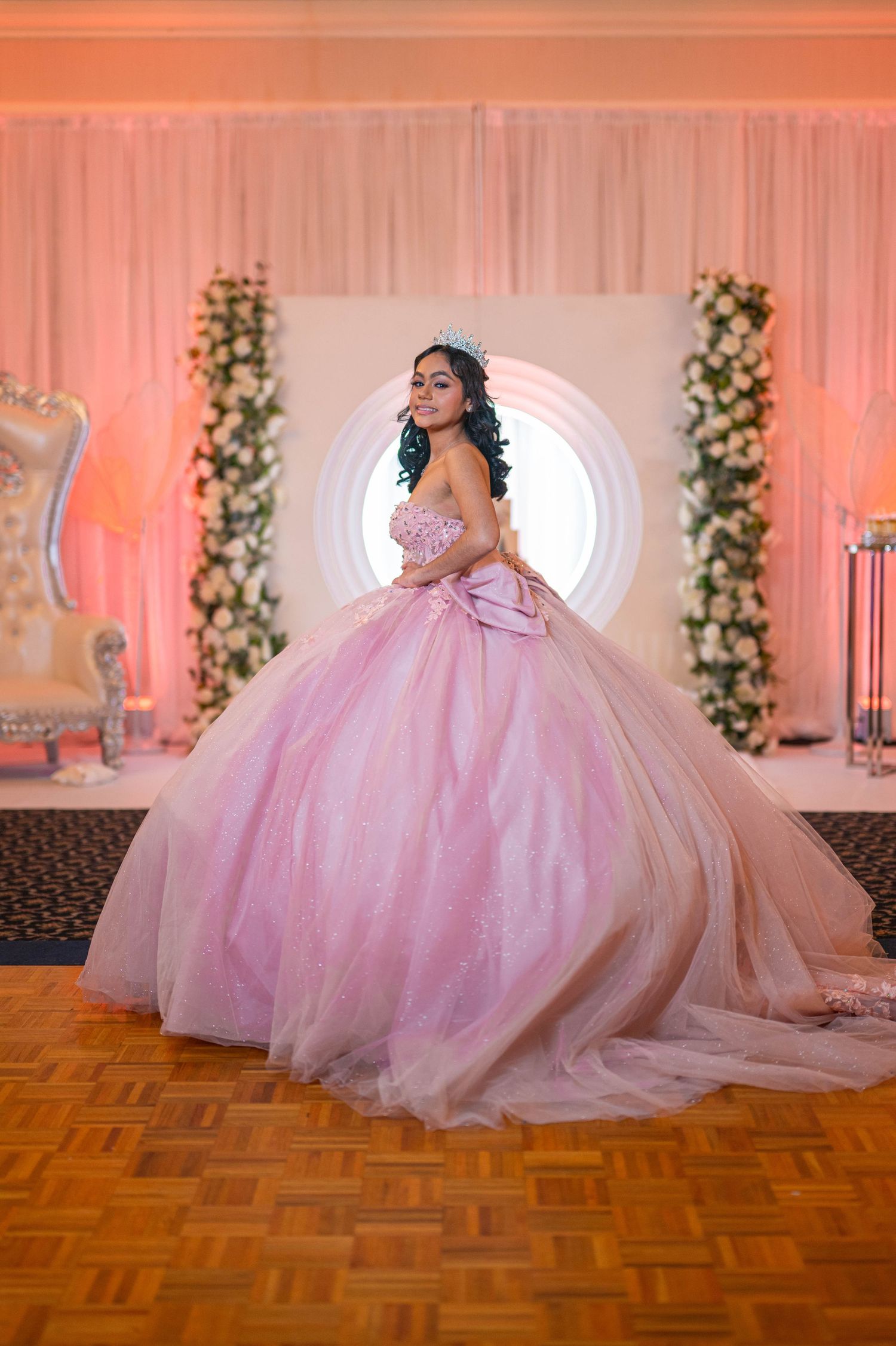 Young woman in pink ball gown quinceañera dress poses elegantly in decorated ballroom with floral backdrop.