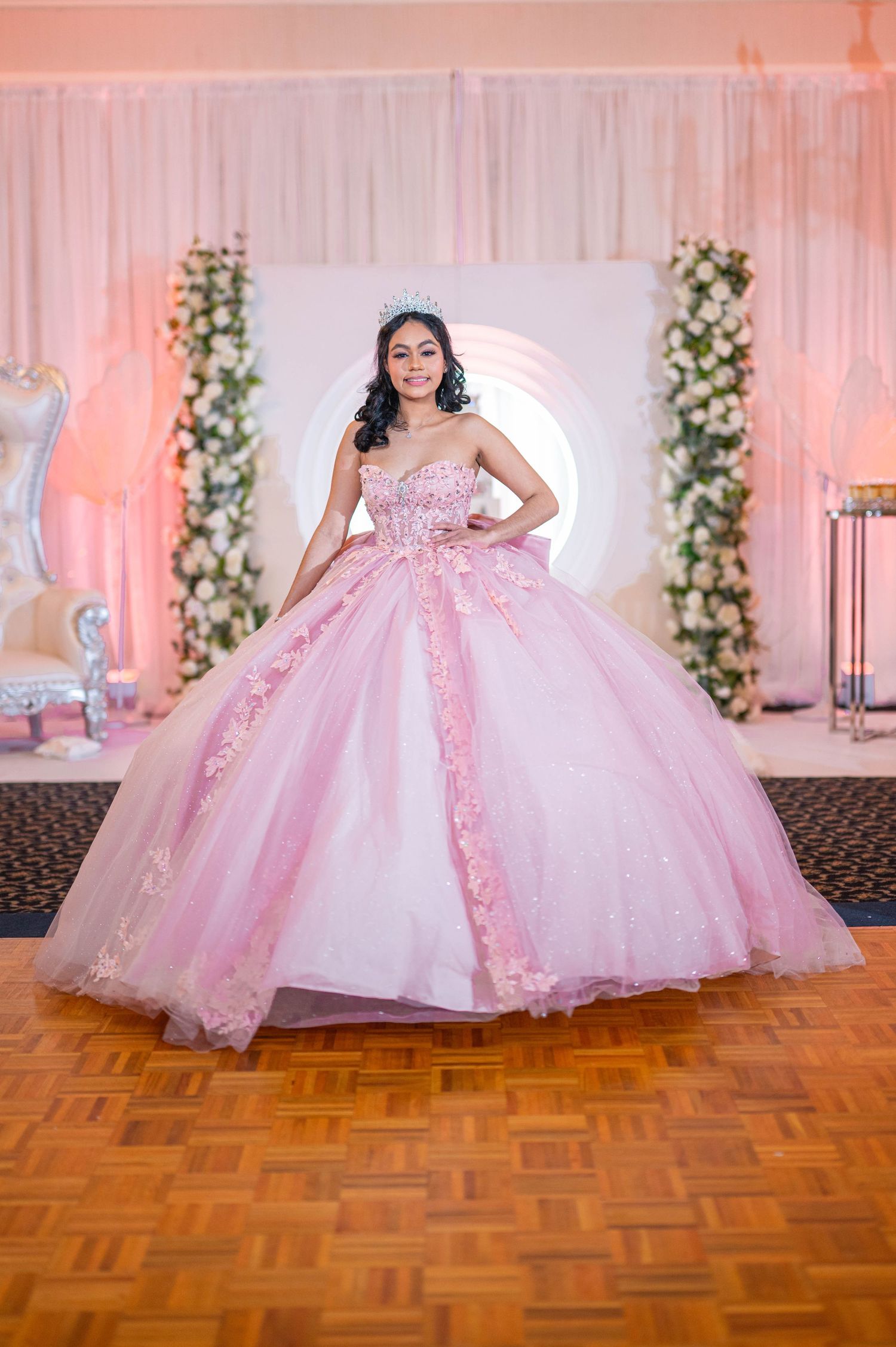Young woman in a pink ballgown quinceañera dress with floral details poses on a dance floor.