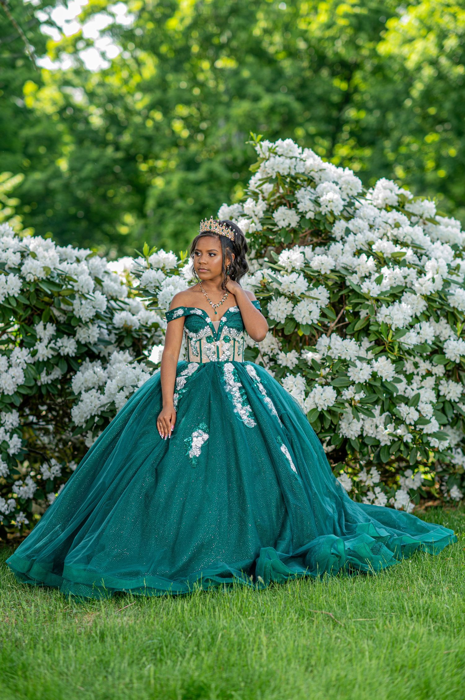 Elegant off-shoulder emerald gown with white lace details set against flowering bushes in a garden.