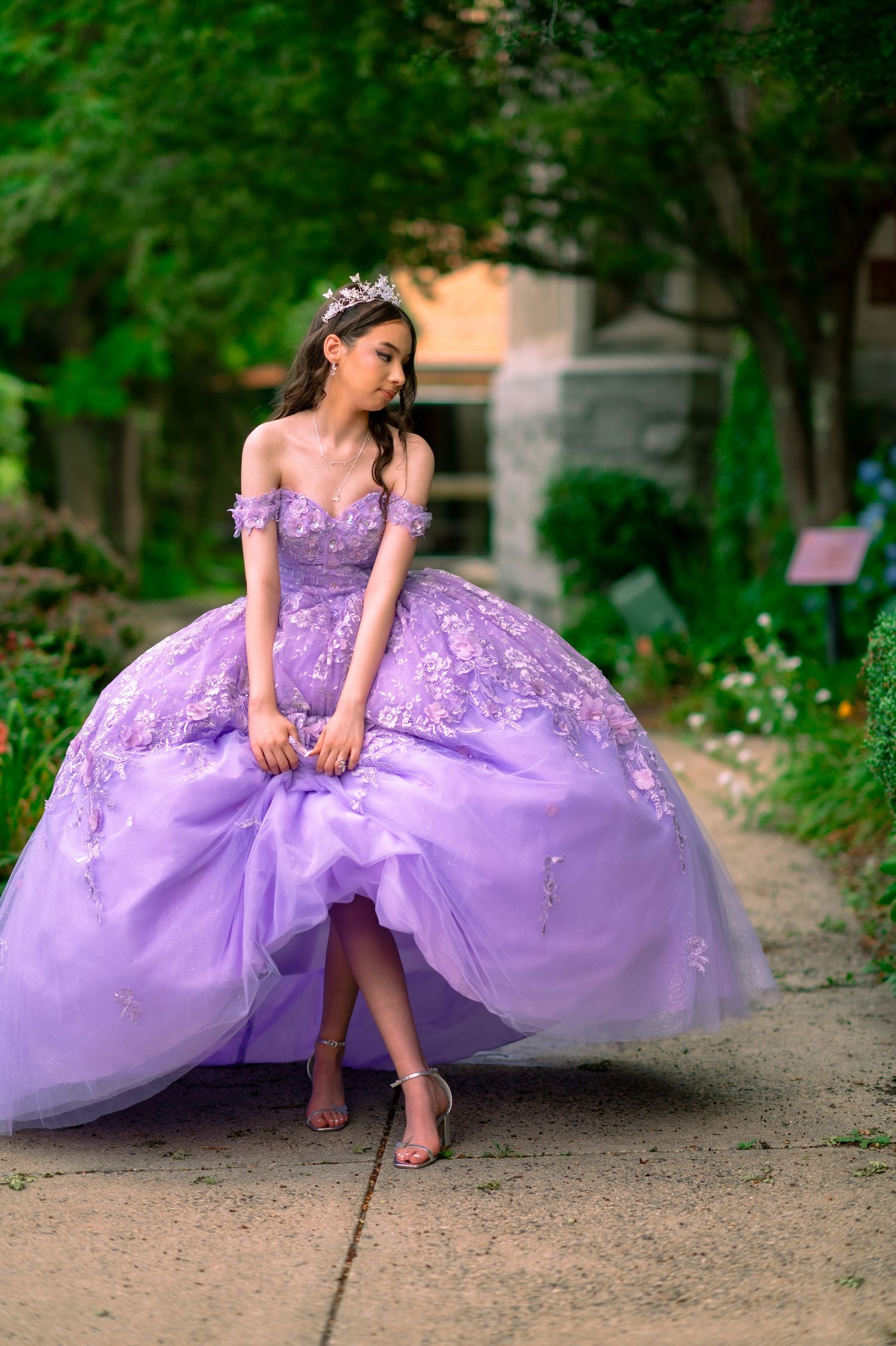 Young woman in a lavender ball gown with floral embroidery and crown poses gracefully outdoors.