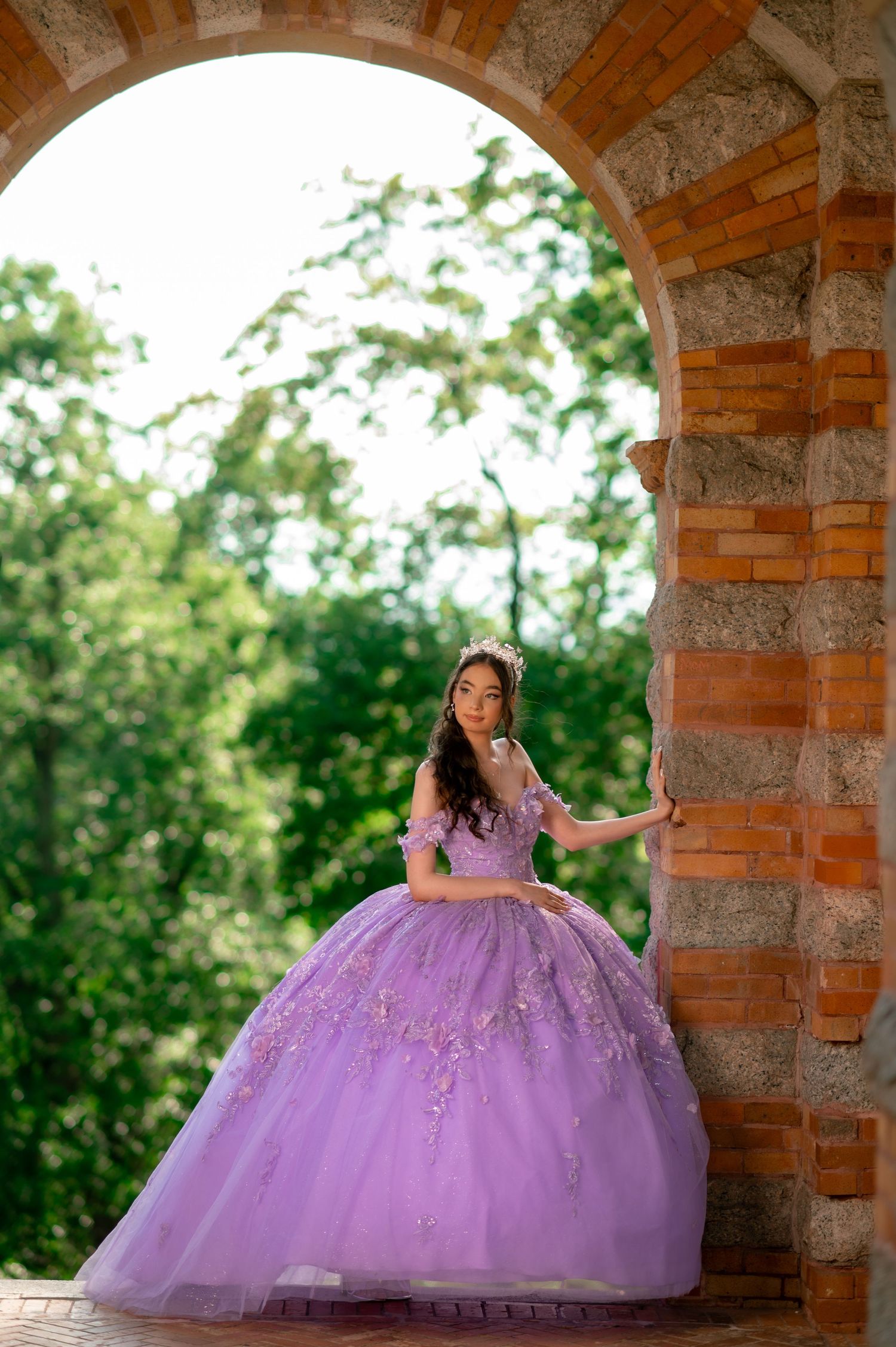 Young woman in lavender quinceañera ball gown with floral embroidery poses under stone arch outdoors.
