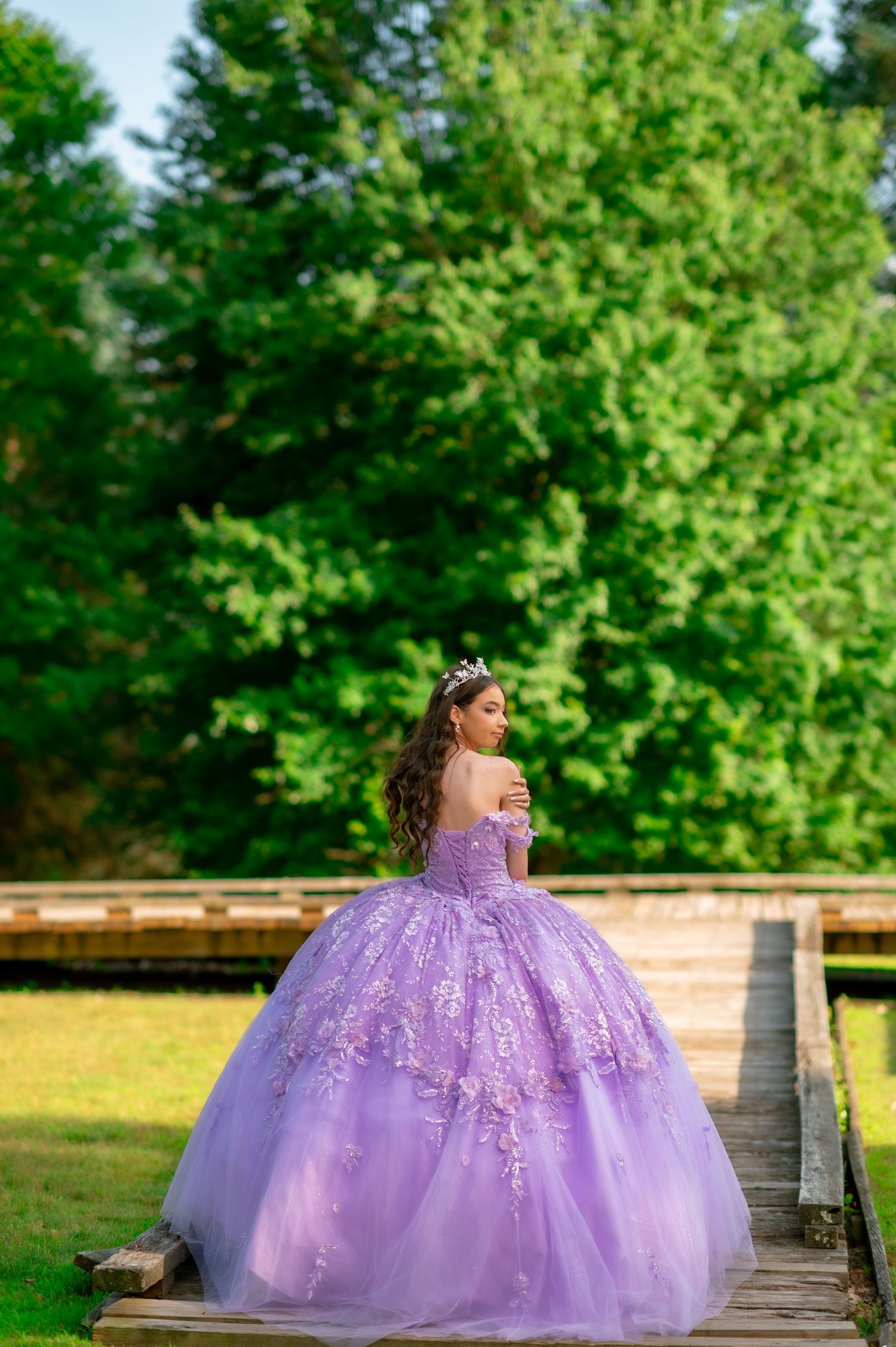 Young woman in lavender ball gown with floral embroidery and tiara poses on wooden bridge surrounded by lush green trees.