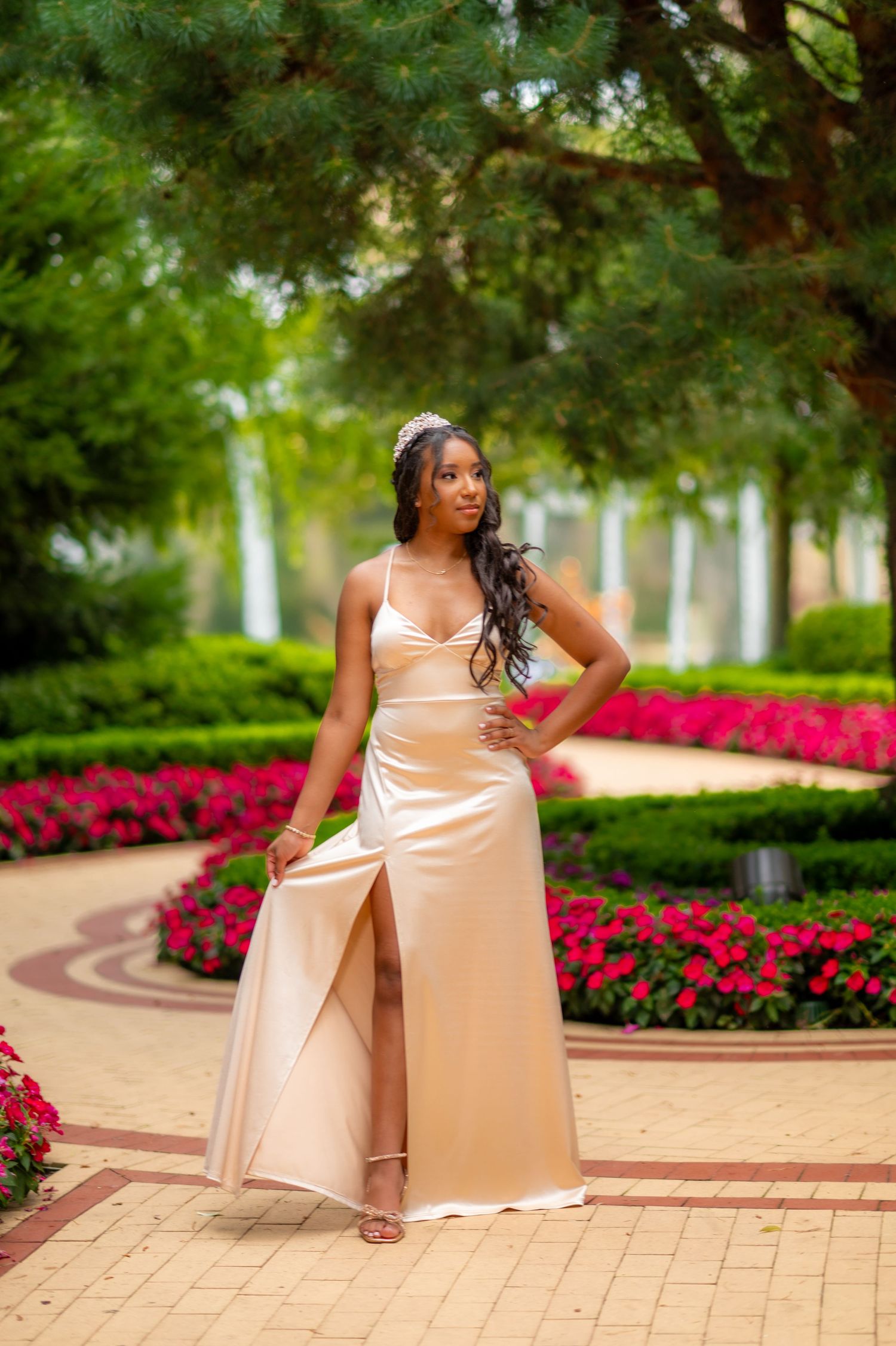 Young woman in elegant gold satin gown with slit poses in a lush garden with red flowers and green hedges.