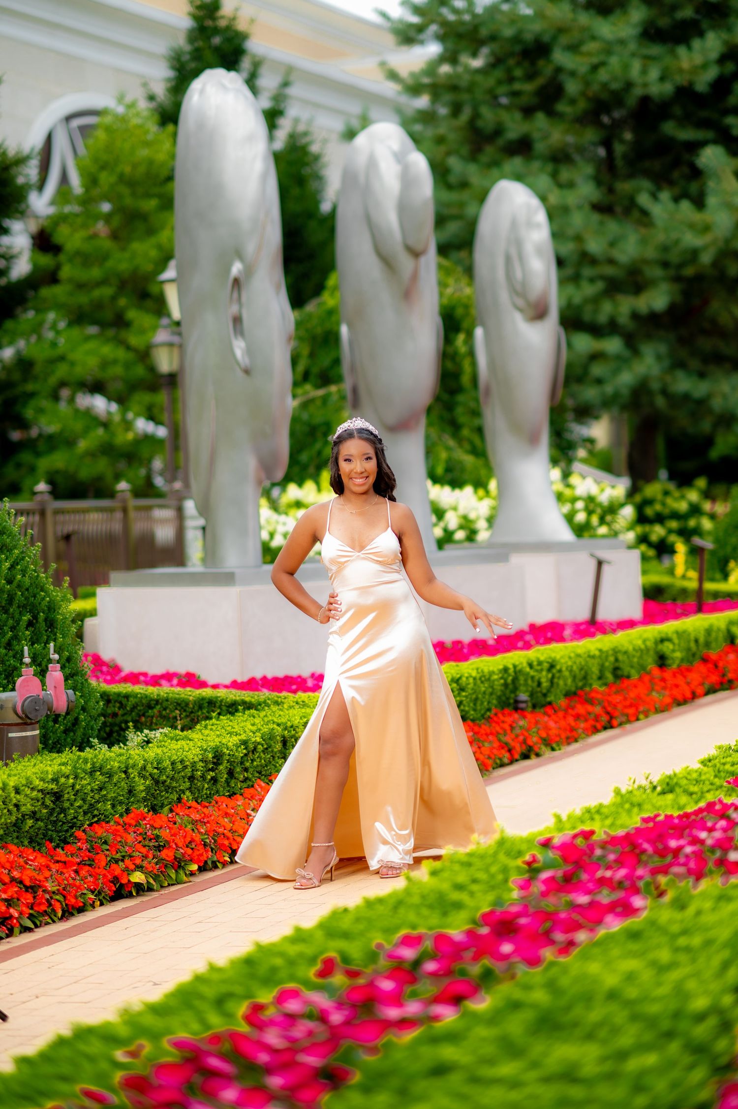 Young woman in elegant gold satin gown posing in a vibrant garden with sculpted statues and colorful flowers.