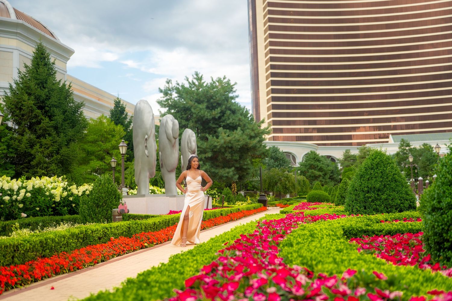 Woman in elegant gown posing in vibrant resort garden with elephant sculptures and colorful flowers.