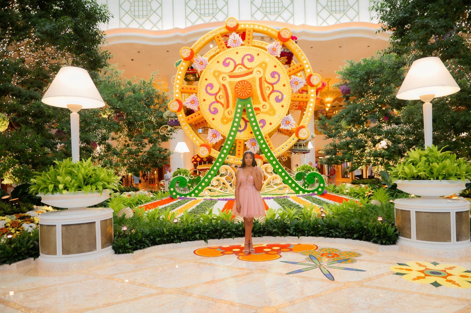 Woman in pink dress posing before a vibrant floral ferris wheel display in an elegant garden atrium.