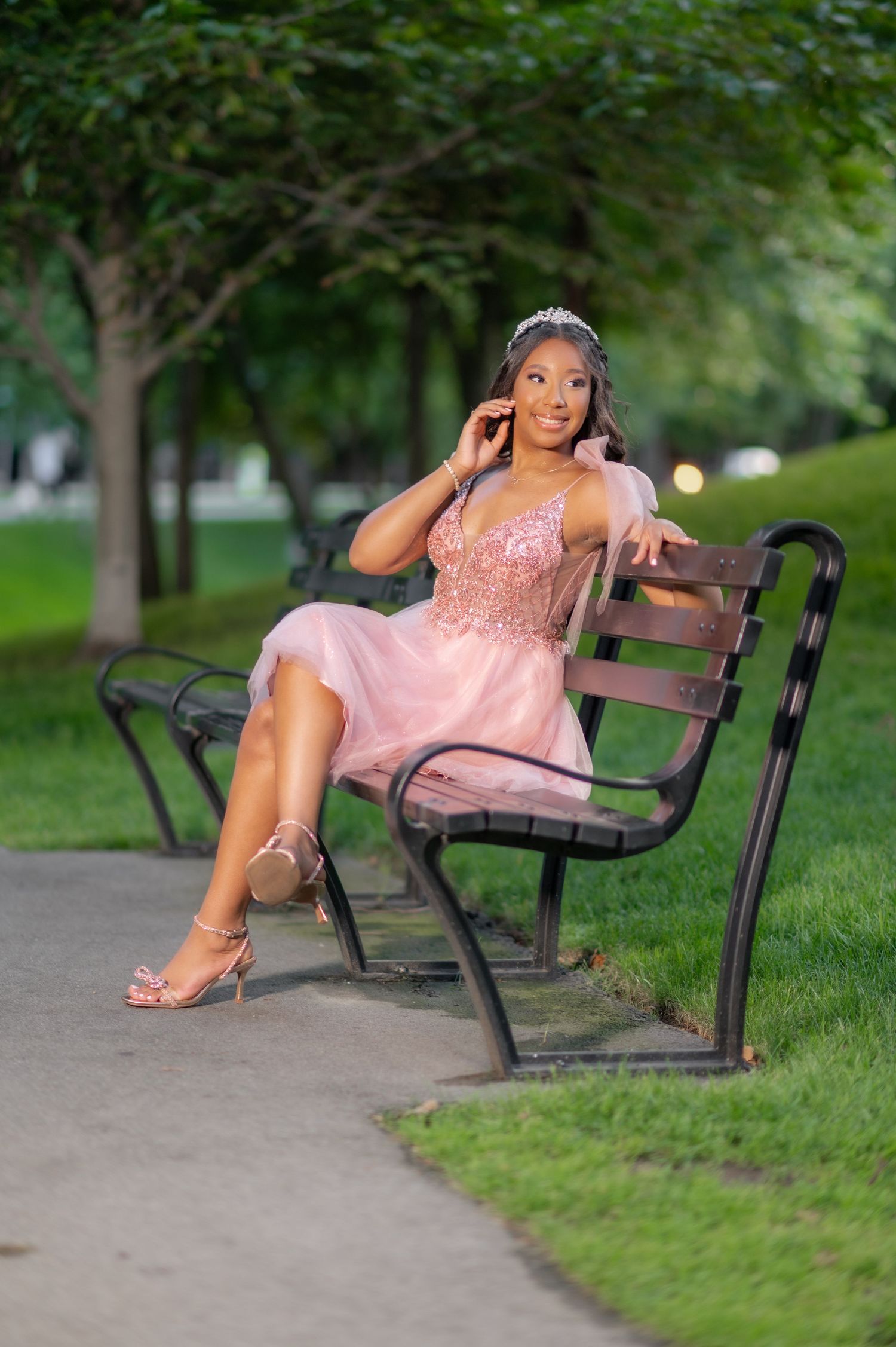 Young woman in pink sequin dress and tiara sits gracefully on park bench surrounded by lush green trees.