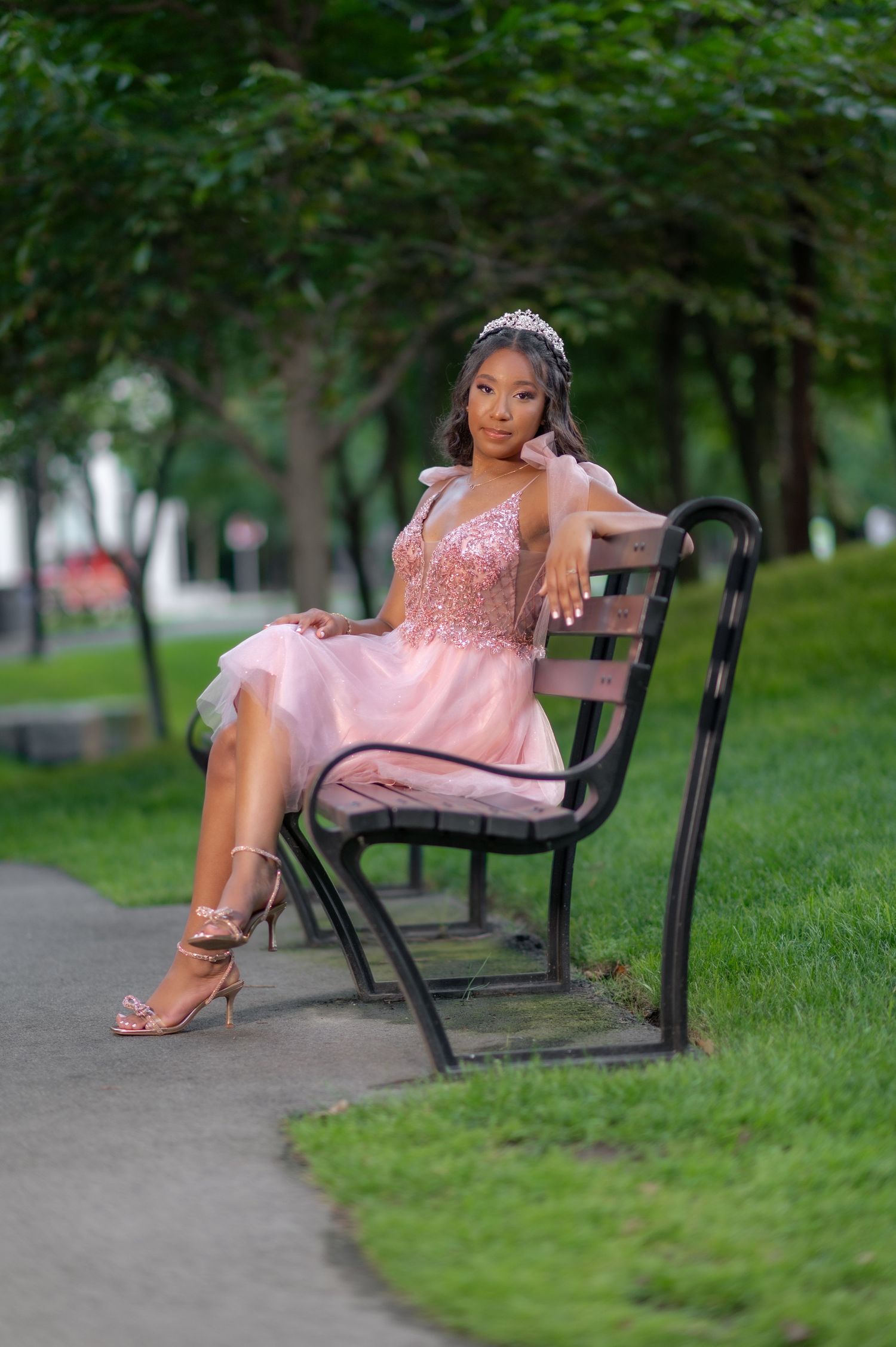 Young woman in pink sequin dress and tiara sits elegantly on a park bench surrounded by lush green trees.