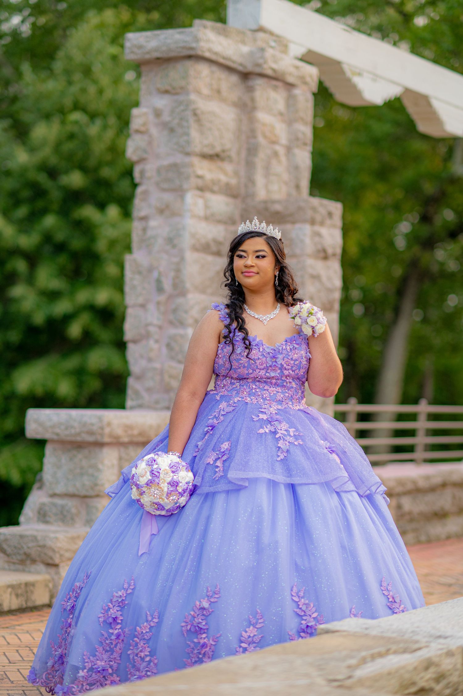 Young woman in lavender quinceañera ball gown with floral appliques, tiara, and bouquet poses outdoors.