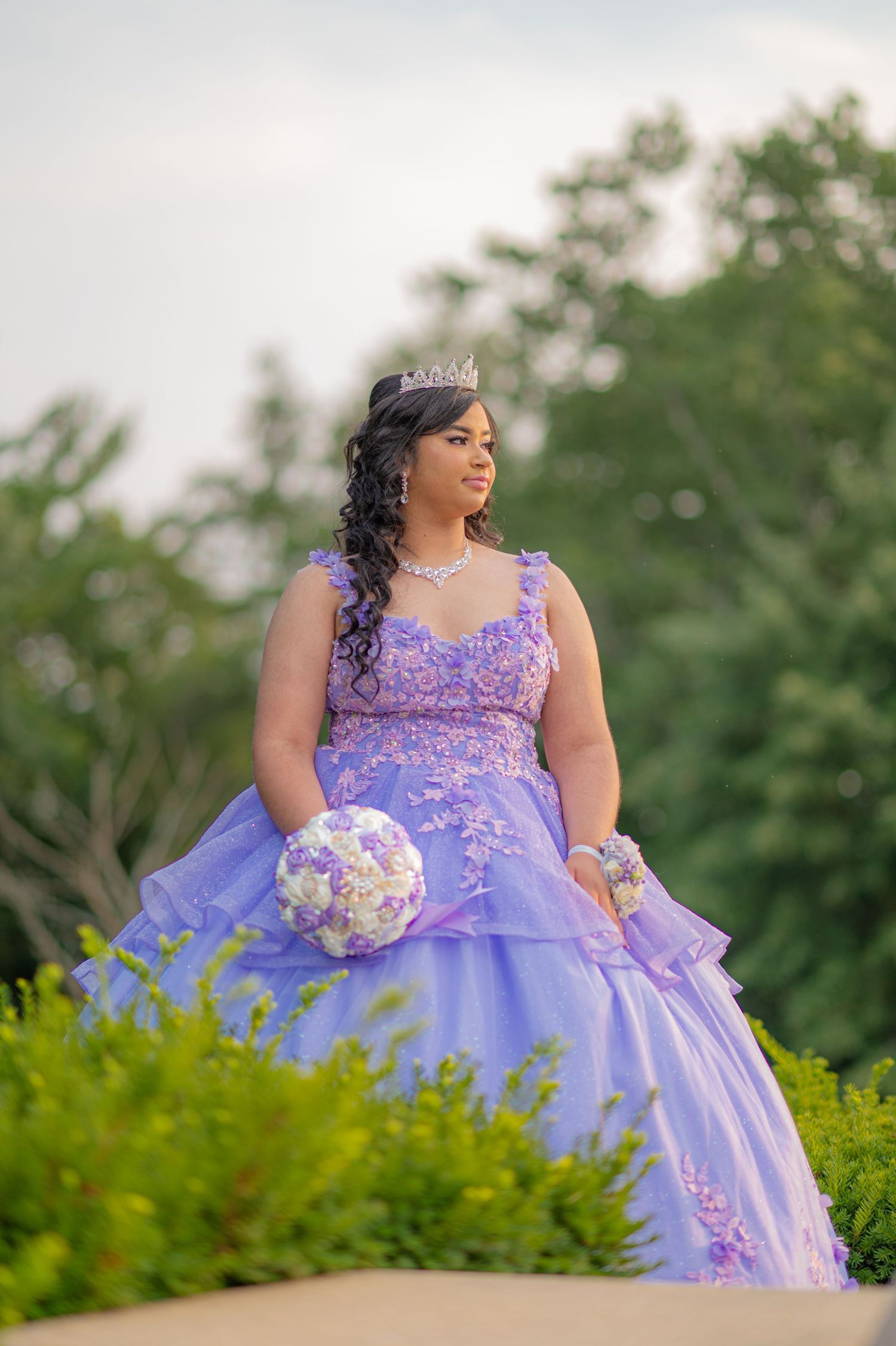 Young woman in lavender quinceañera ball gown with tiara holds floral bouquet outdoors among lush green trees.