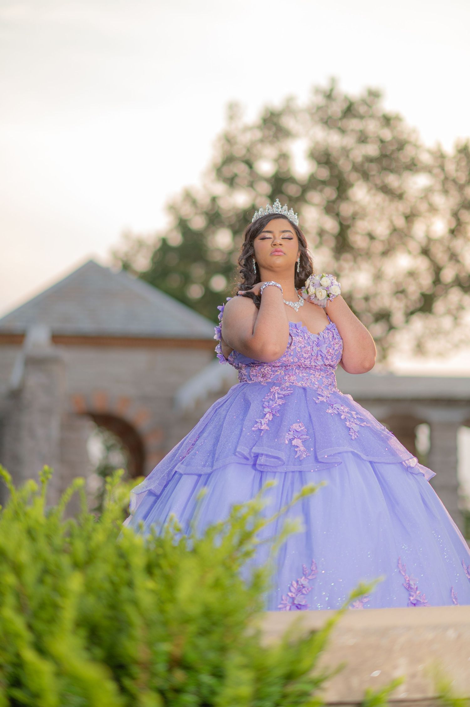 Young woman in lavender quinceañera ball gown with tiara poses outdoors at golden hour.