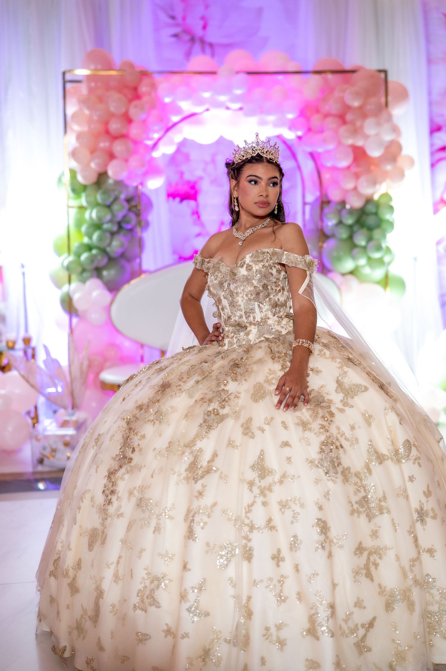 Young woman in gold quinceañera ball gown with crown poses at elegant pink and green balloon decorated celebration.