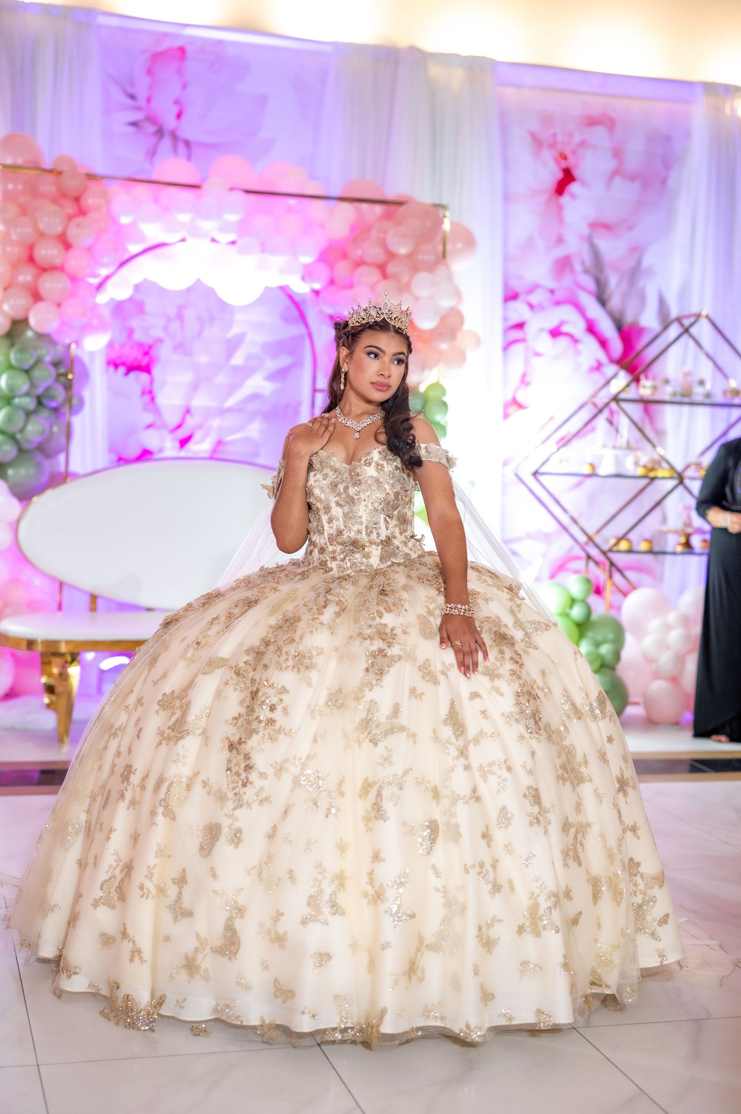Young woman in gold quinceañera ballgown with tiara poses at elegant celebration with balloon decorations.