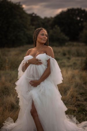gravid kvinna står på en äng. hon har en vit tyllklänning på sig och håller om sin gravidmage