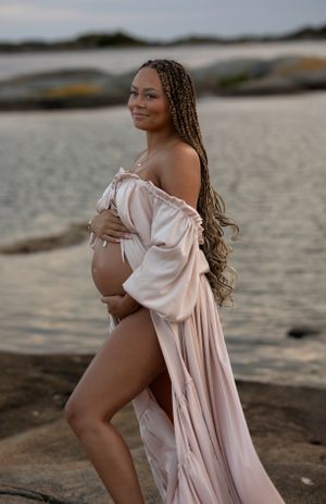 kvinna står på en klippa iklädd en rosa klänning. hon håller om sinn gravidmage och kollar bortåt och ler. i bakgrunden syns havet.