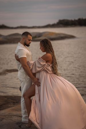 en gravid kvinna och en man står emot varann och håller om varann. de står på en klippa vid havet vid solnedgången och kollar neråt.