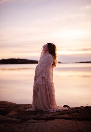gravid kvinna iklädd en ljus klänning på en klippa i solnedgången. hon håller ena handen i håret och andra på sin gravidmage