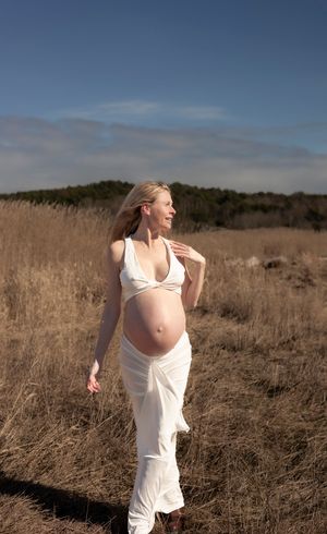gravid kvinna står på en äng. på sig har hon ett vitt set. hon kollar bortåt och ler. ena handen har hon på sin axel.