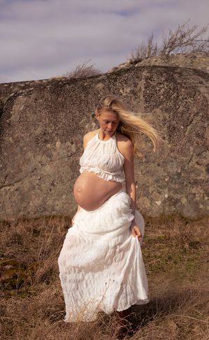 gravid kvinna står på en gräsplätt och bakom henne är en klippa. hon har ett vitt set på sig.