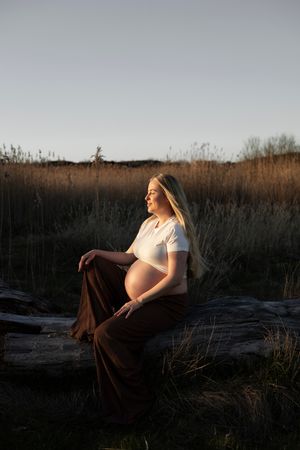 gravid kvinna sitter ned på en stock och har solens gyllene sken på sig. på sig har hon bruna byxor och en vit t-shirt.