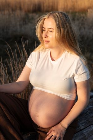gravid kvinna sitter ner och har solens gyllene sken på sig. på sig har hon bruna byxor och en vit t-shirt.