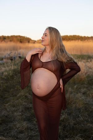 en gravid kvinna står på en äng och har en brun klänning ned ett hål för magen. hon har handen på axeln och kollar bortåt.