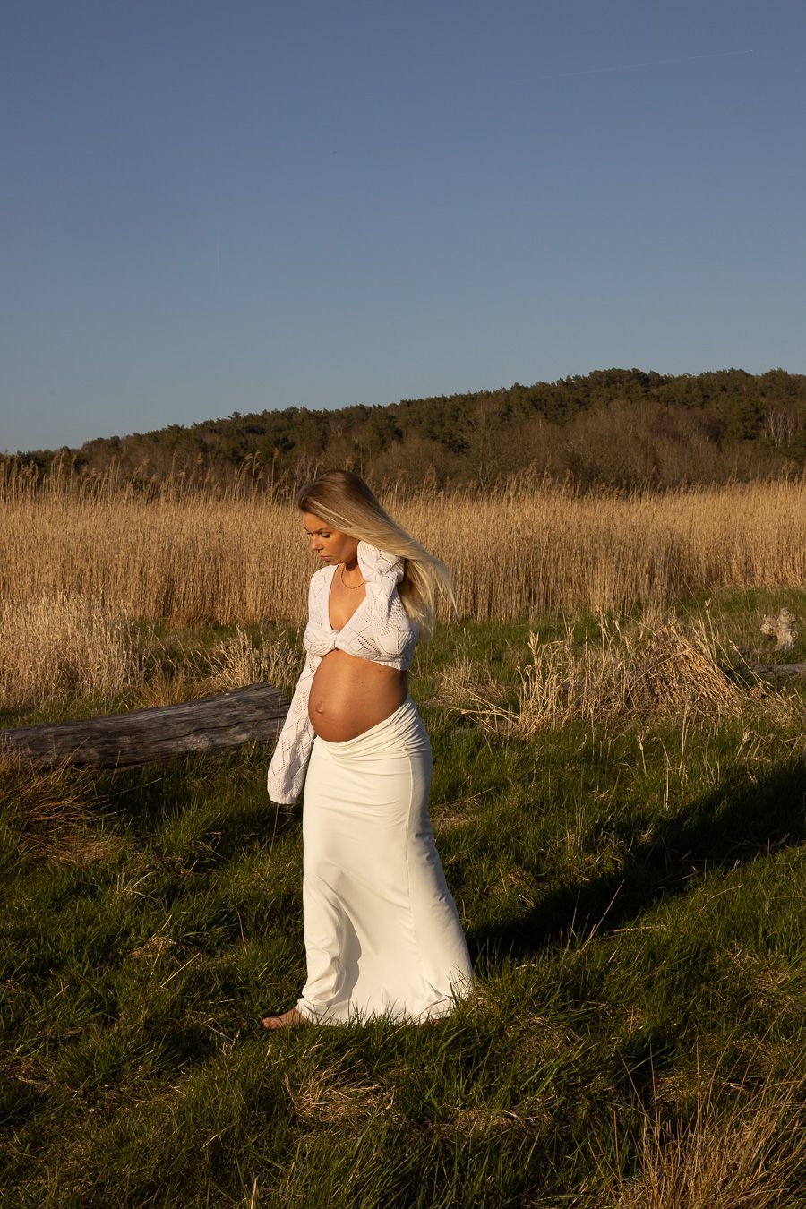 gravid kvinna går på en åker, himmelen är blå i bakgrunden. hon har ett vitt set på sig och kollar ner i marken.