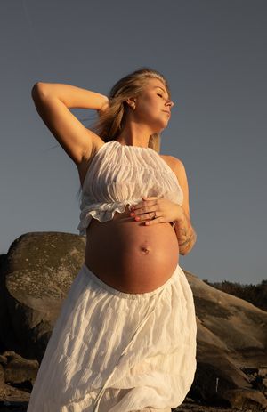 gravid kvinna i fokus med en klippa i bakgrunden. solens gyllene sken lyser på henne. hon har ett vitt set på sig.