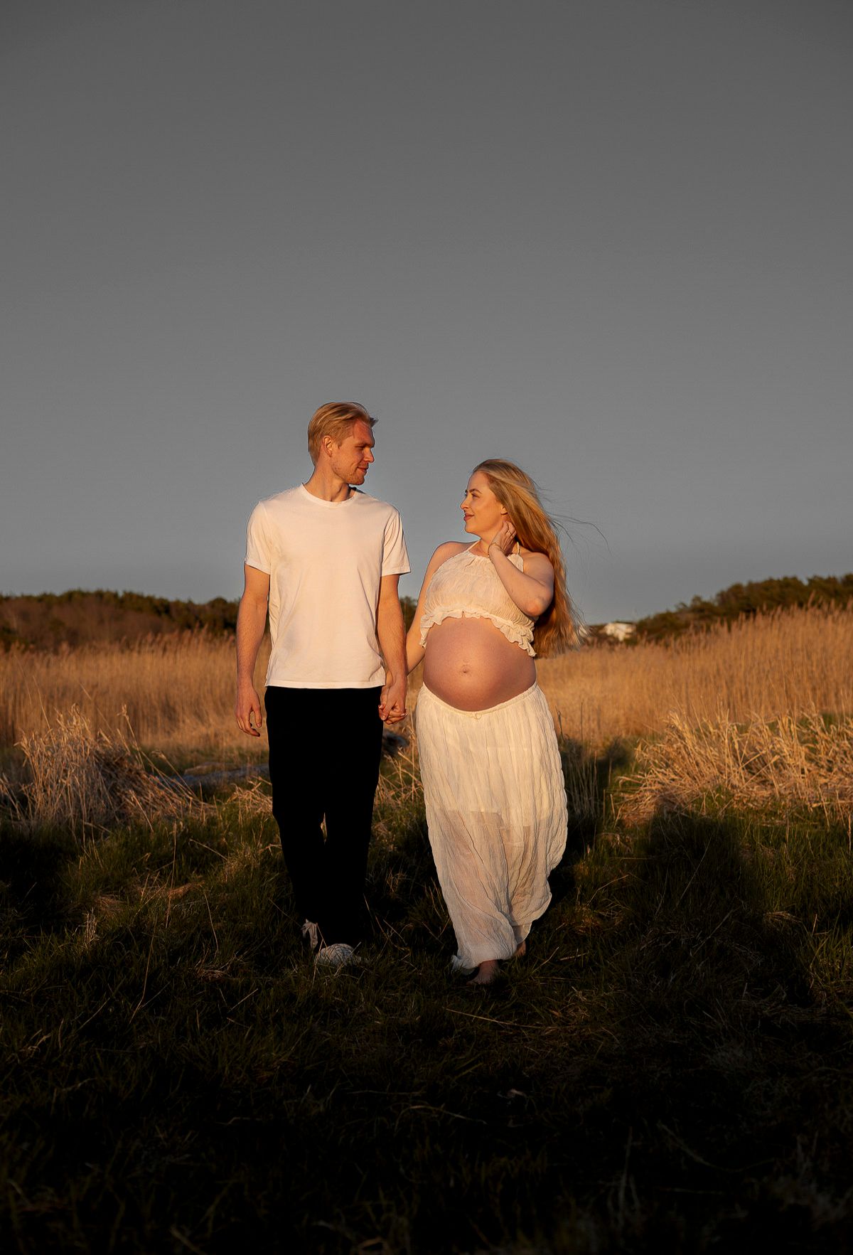 en man och en gravid kvinna går i solens gyllene ljus på en äng och håller varandra i händerna. de kollar mot varandra.