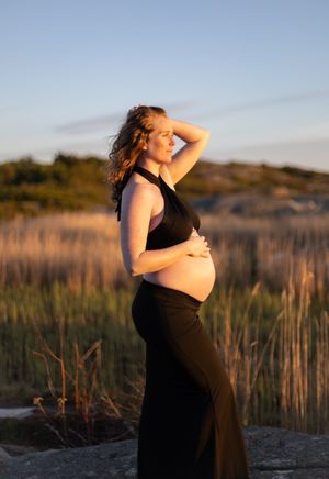 en gravid kvinna som står och kollar utåt. hon har en svart topp och kjol på sig och håller på sin gravidmage.