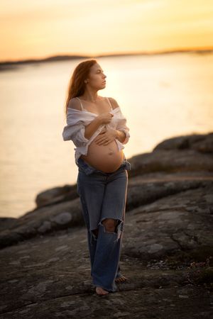 gravidfotografering i solnedgången vid havet. kvinnan står på en klippa och är kläd i jeans och en vit tröja och är barfota.