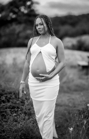 bilden består av en gravid kvinna som står på en äng klädd i en vit topp och en vit kjol. hon håller på sin gravidmage. bilden är svartvit.