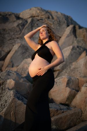 i bakgrunden syns klippor. en gravid kvinna som står och kollar utåt. hon har en svart topp och kjol på sig och håller på sin gravidmage