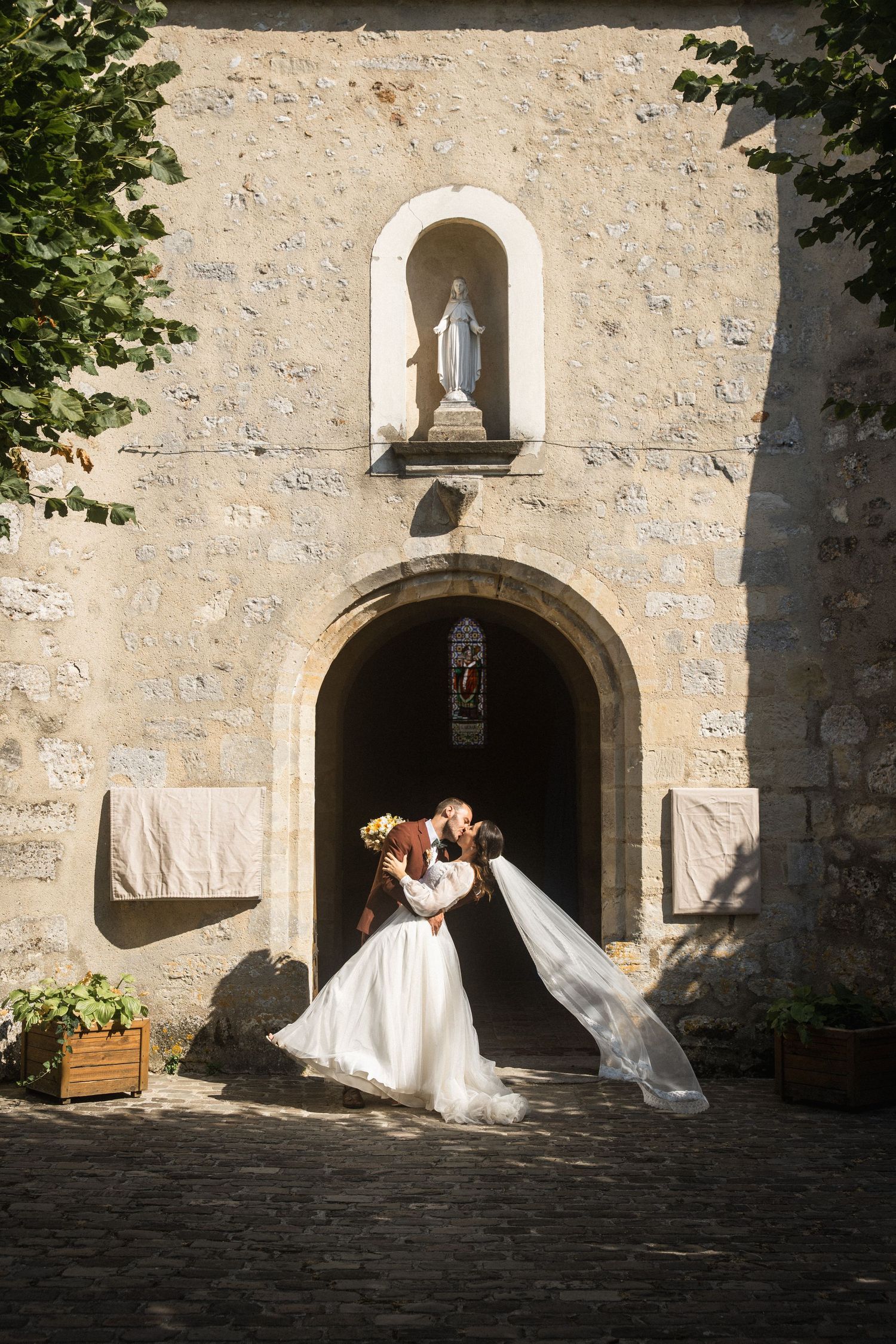 Photo d'un couple de mariés à la sortie d'une magnifique église, s'embrassant sous un soleil d'été dans leur tenues élégantes