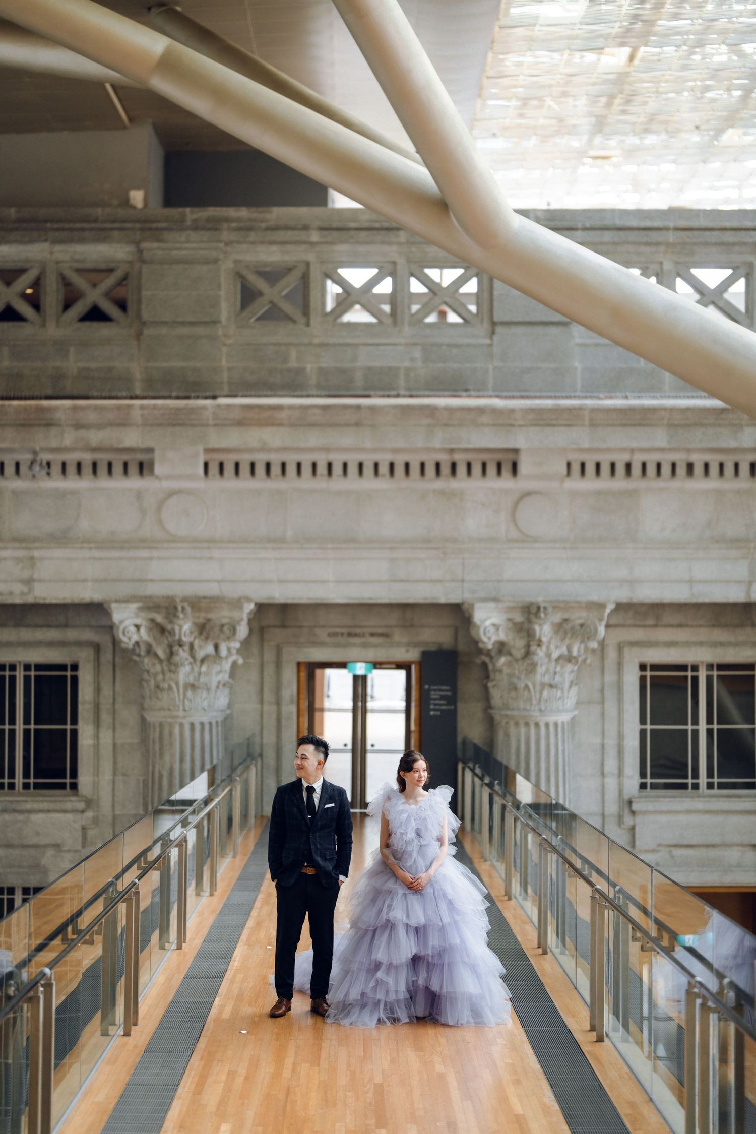 A couple walks along an elegant indoor corridor with classical architectural details and wooden flooring.