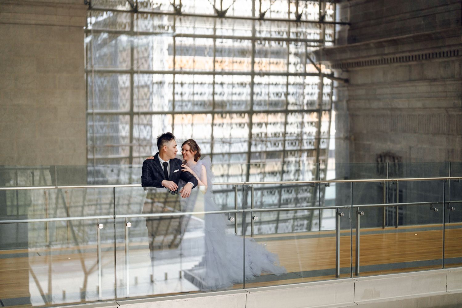 A couple embraces in a bright indoor gallery with large windows and city views.
