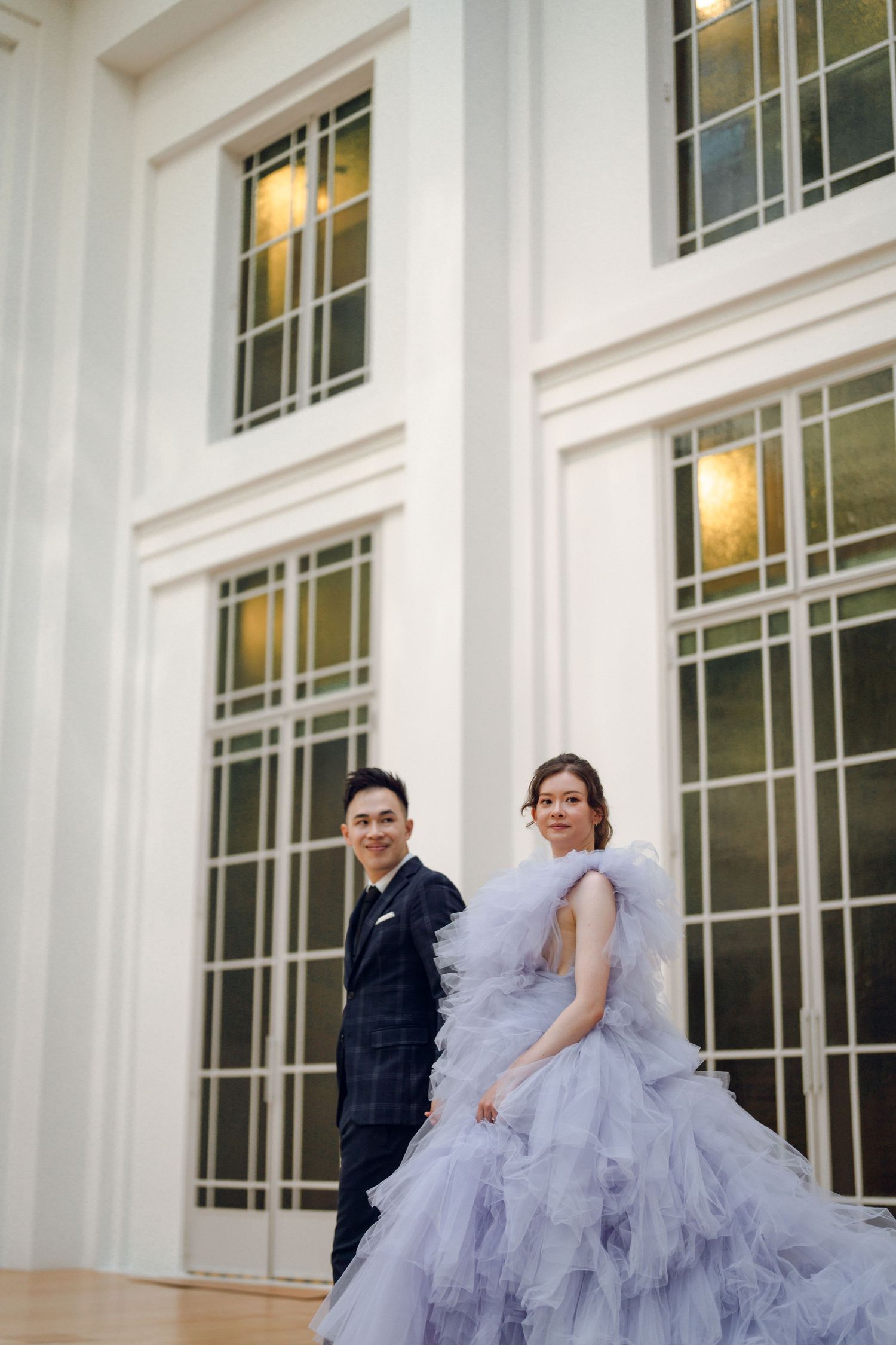 A couple poses against white architectural columns and windows in an elegant indoor setting.